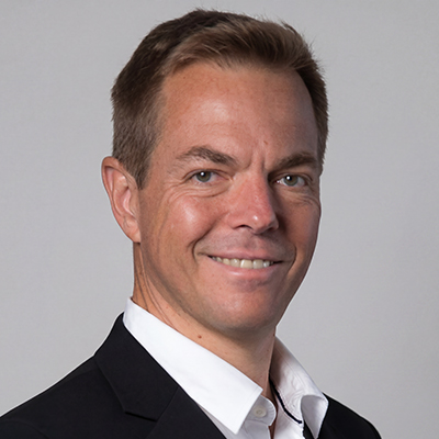 A man with short light brown hair wearing a black blazer and white collared shirt smiles at the camera against a plain light grey background.