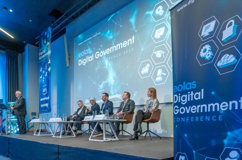 A panel of five people sits onstage at the Eolas Digital Government Conference 2025, with a speaker at a lectern. Blue digital-themed graphics and banners appear behind them, displaying conference branding and technology icons.