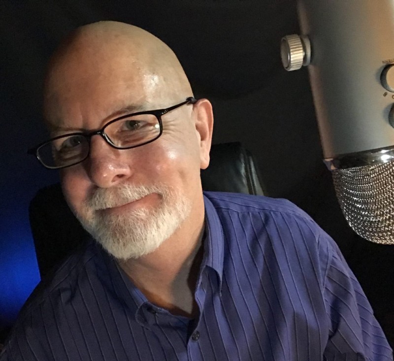 Un homme chauve souriant avec des lunettes, une barbe blanche et une chemise à rayures bleues est assis devant un grand microphone argenté dans une pièce faiblement éclairée.