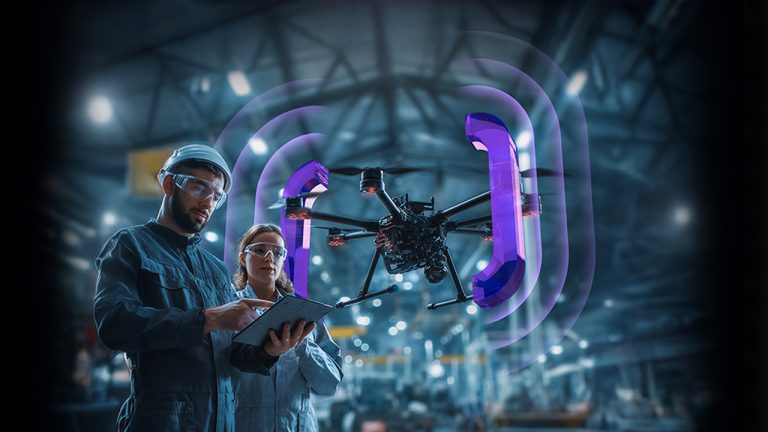 Two engineers in protective gear operate a tablet whilst inspecting a large drone with glowing purple signal arcs, inside a high-tech industrial facility. The background is blurred, emphasising the drone and engineers.