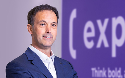 A man in a blue suit stands smiling in front of a purple background with partial white text that reads "( exp" and "Think bold.