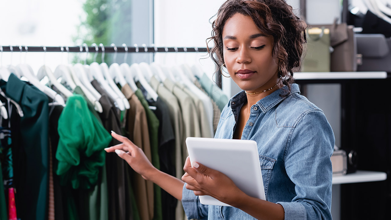 A woman in a denim shirt holds a tablet and uses her other hand to sort through clothes on hangers in a clothes shop. Shelves and rails of clothes are visible in the background.