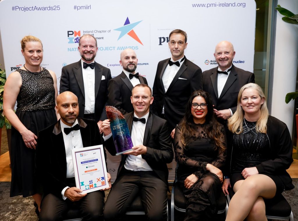 Eight people dressed smartly, some sitting and some standing, pose for a photo at an awards event. One man holds an award trophy and another holds a framed certificate. Event banners are visible in the background.