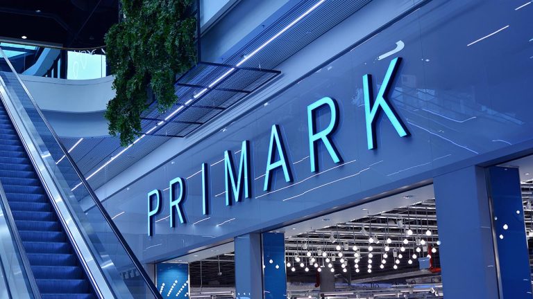 Blue neon “PRIMARK” sign on the outside of a modern shop next to an escalator, with plants hanging above and bright interior lights visible through the glass.