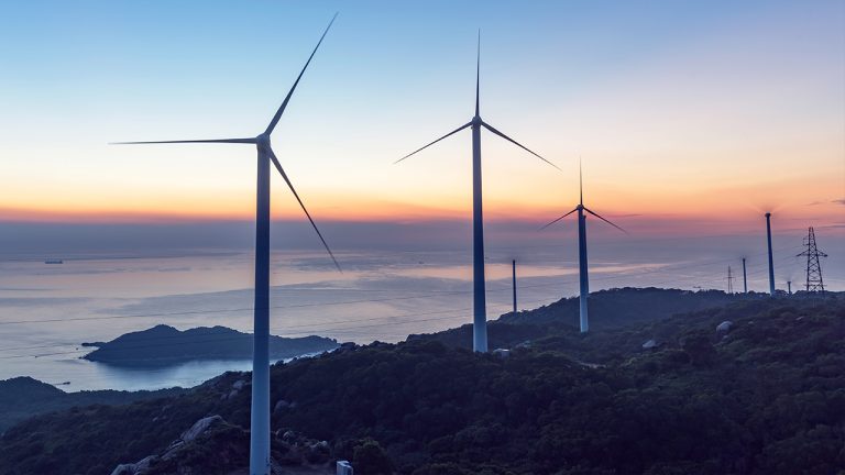 Three large wind turbines stand on a hilly landscape overlooking the sea at sunset, with an orange and blue sky and a distant island visible in the background. Power lines and a ship can also be seen.