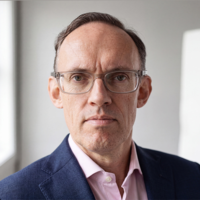 A middle-aged man with short brown hair, wearing clear-framed glasses, a navy blue suit, and a light pink dress shirt, looks directly at the camera in a bright, softly lit room.