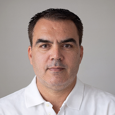 A man with short dark hair and stubble, wearing a white collared shirt, looks directly at the camera against a plain, light grey background.