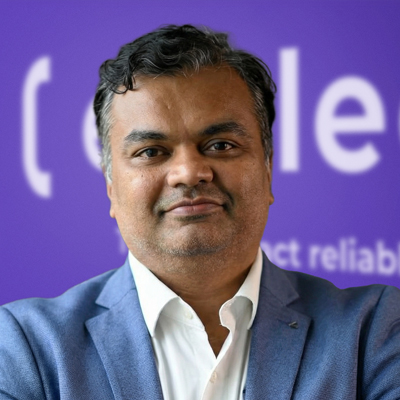 A man with dark hair wearing a blue suit jacket and white shirt stands in front of a purple background with partial white text. He is facing the camera and smiling slightly.