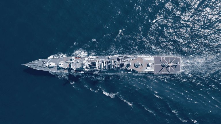 Aerial view of a large navy ship sailing through deep blue water, leaving a wake behind. The ship features a helipad at the rear with a helicopter outline visible. Sunlight reflects off the water.