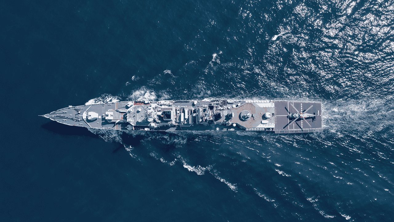 Aerial view of a large navy ship sailing through deep blue water, leaving a wake behind. The ship features a helipad at the rear with a helicopter outline visible. Sunlight reflects off the water.