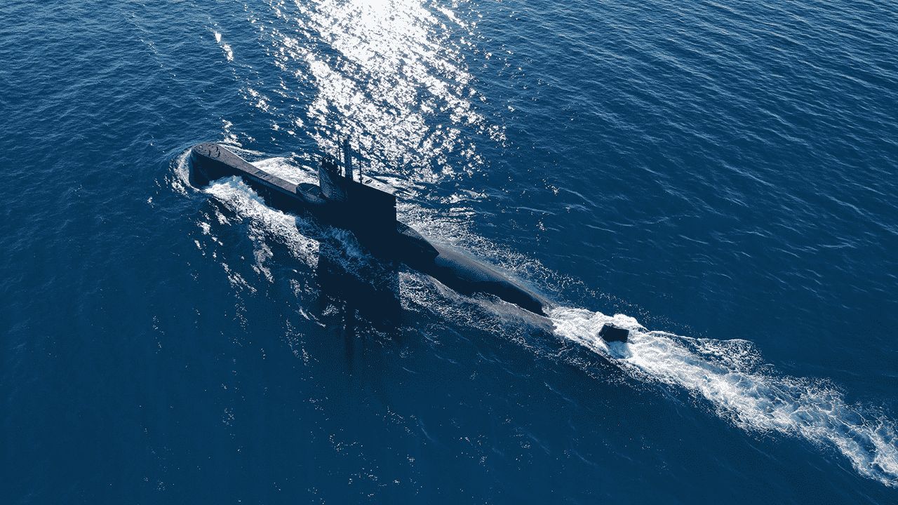 A submarine cruises on the surface of calm blue ocean water, creating a gentle wake behind it under bright sunlight.
