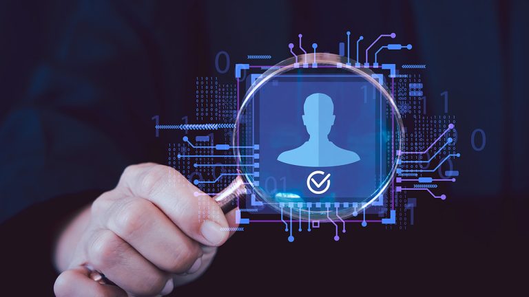 A hand holds a magnifying glass over a digital silhouette of a person with a tick, surrounded by circuit and binary designs, symbolising identity verification and cybersecurity.