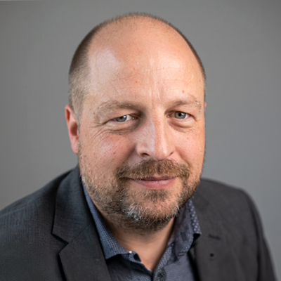 A middle-aged man with a bald head, light beard, and blue eyes is smiling slightly. He is wearing a grey blazer over a dark shirt and is posed in front of a plain grey background.
