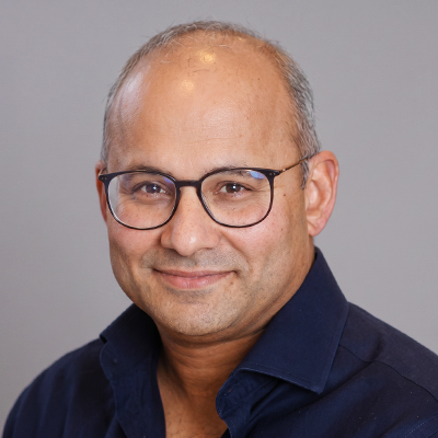 A middle-aged man with glasses, short hair, and a receding hairline, wearing a navy blue collared shirt, smiling softly against a plain grey background.