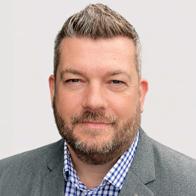 A man with short, grey hair and a beard, wearing a grey blazer over a blue and white chequered shirt, smiling softly against a plain light background.