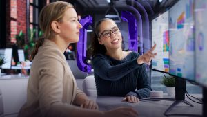 Two women sit at a desk in an office, looking at a computer monitor displaying colourful charts and graphs. One woman points at the screen whilst explaining something to the other.