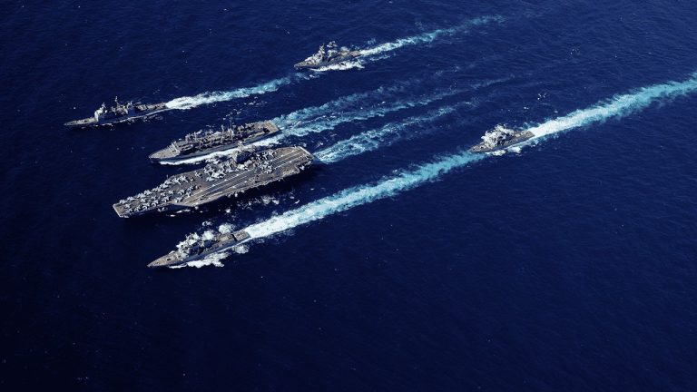 Five navy ships, including an aircraft carrier, sail in formation across deep blue ocean water, leaving white trails behind them in a coordinated pattern.