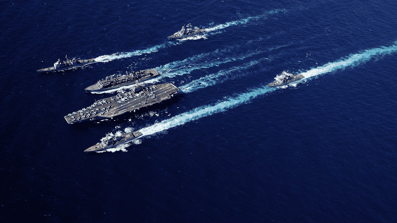 Five navy ships, including an aircraft carrier, sail in formation across deep blue ocean water, leaving white trails behind them in a coordinated pattern.