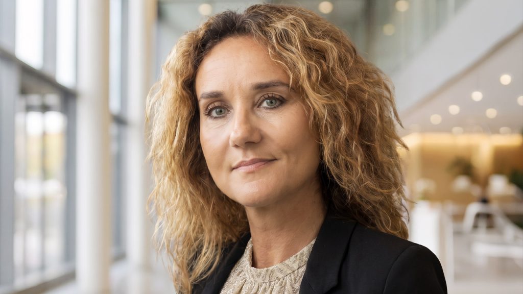 A woman with curly blonde hair and a black blazer stands indoors with large windows and modern decor in the background.
