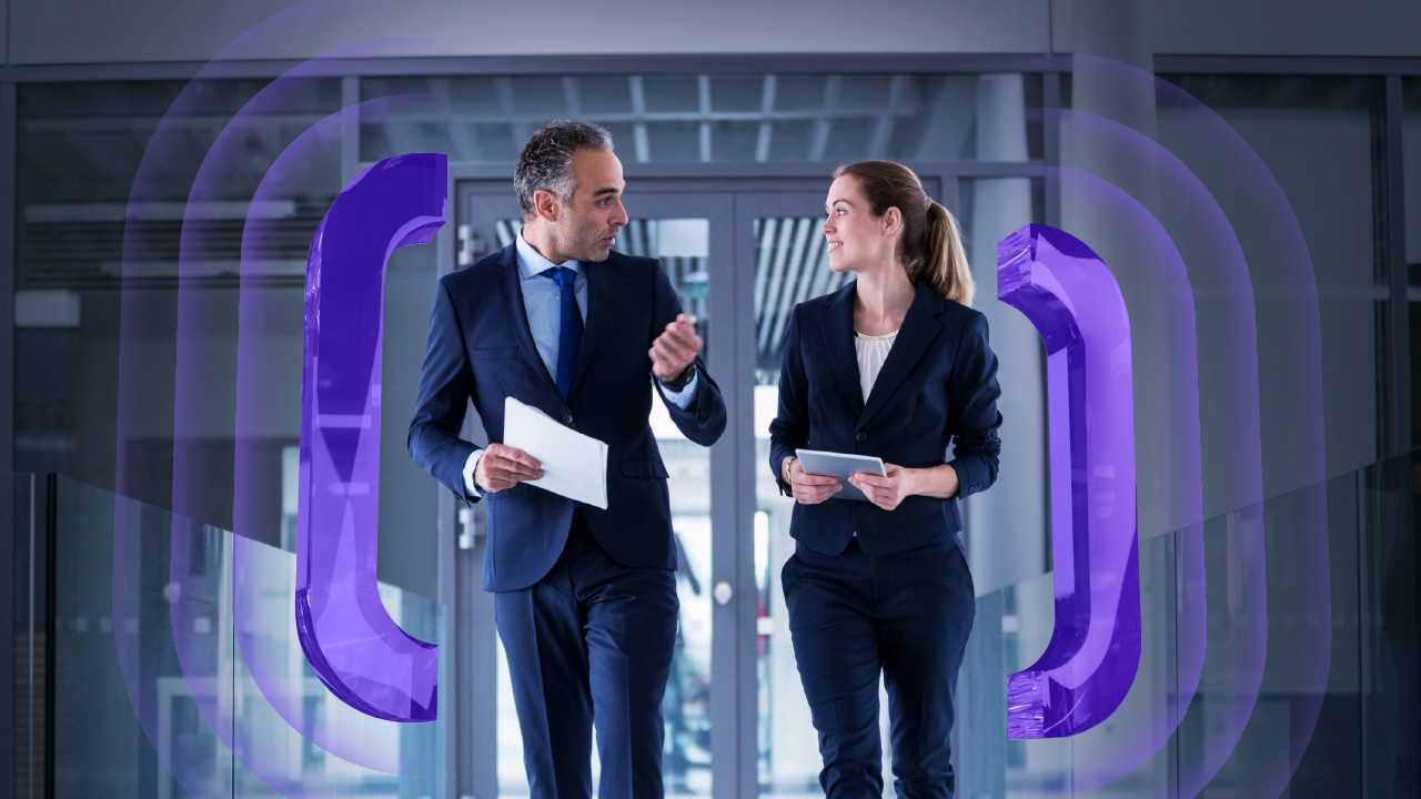 Two business professionals in suits walk down an office corridor, holding documents and talking. Purple abstract parentheses-like shapes are digitally added, framing them on both sides.