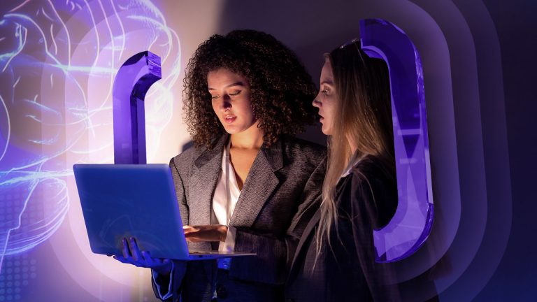 Two women in business attire work on a laptop beside a glowing digital illustration of a brain and large purple quotation marks.