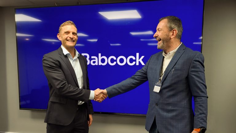 Two men in suits shake hands in front of a digital screen displaying the Babcock logo.
