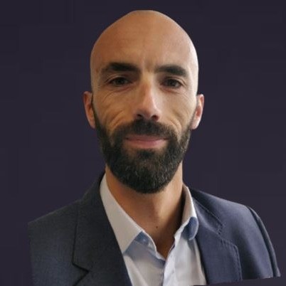 Un homme chauve et barbu, vêtu d'une veste de costume bleue et d'une chemise bleu clair, pose sur un fond uni et sombre.