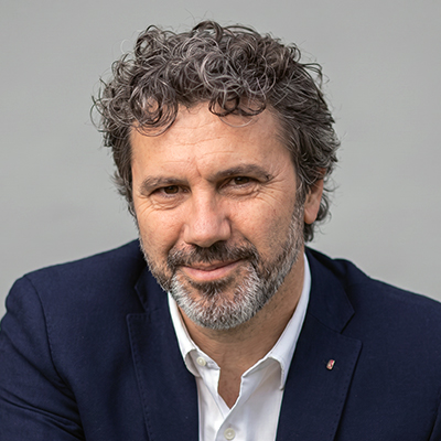 Un homme aux cheveux et à la barbe bouclés, poivre et sel, vêtu d'un blazer bleu marine et d'une chemise blanche, souriant doucement sur un fond uni gris clair.