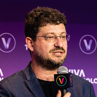 Un homme aux cheveux bouclés et portant des lunettes s'exprime au micro lors d'un événement, debout devant une toile de fond violette sur laquelle figurent l'inscription « VIVA » et des logos circulaires en forme de « V ».