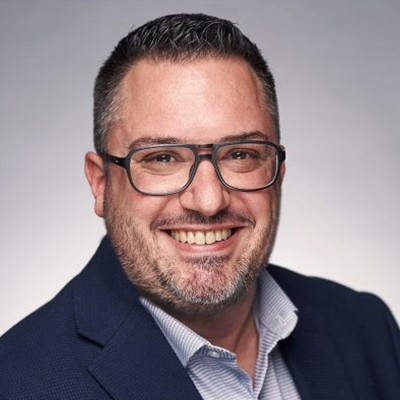 Un homme souriant, aux cheveux courts et foncés, portant une barbe et des lunettes, vêtu d'un blazer bleu marine et d'une chemise rayée, posant devant un fond gris neutre.