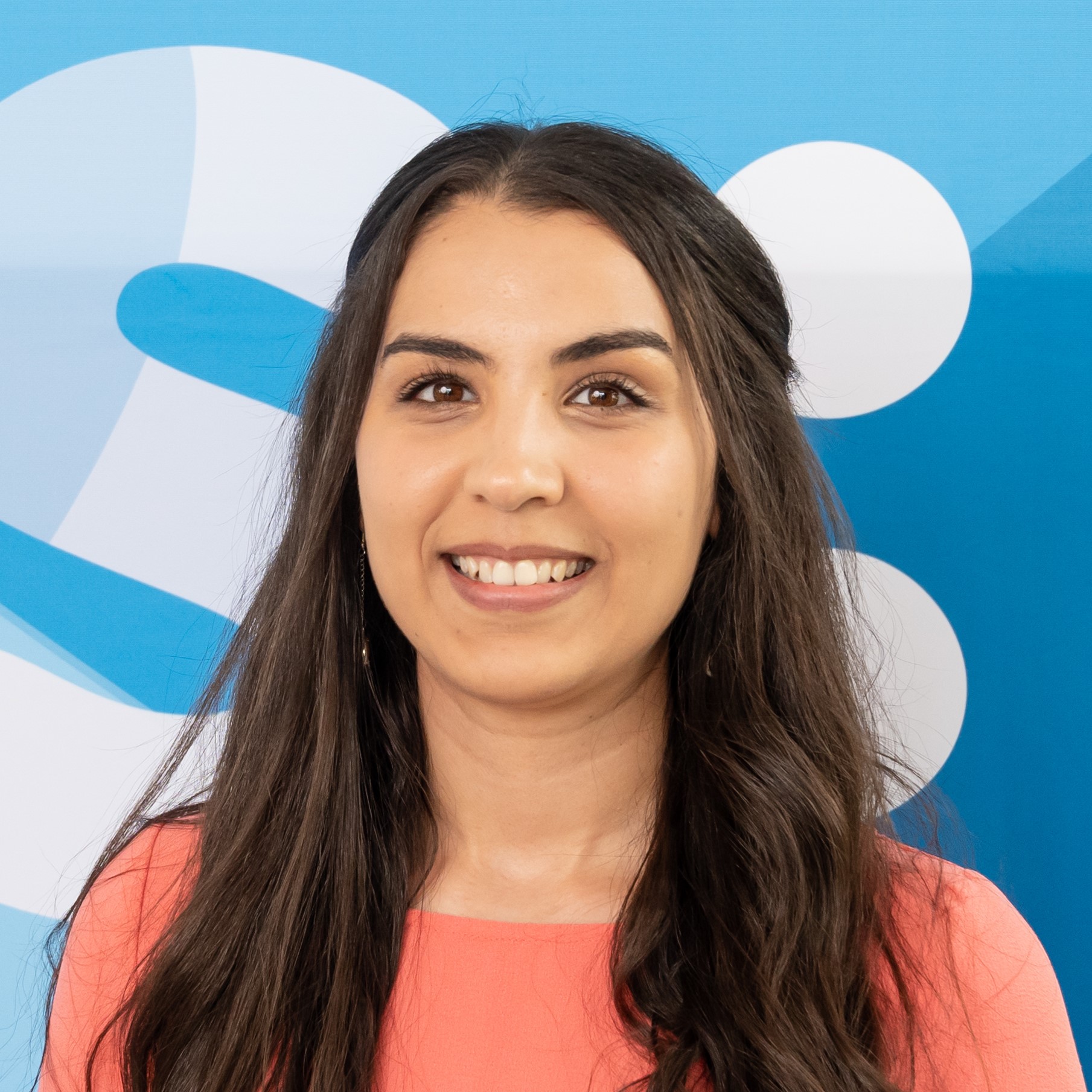 Une jeune femme aux longs cheveux noirs et au haut corail sourit devant un fond abstrait bleu et blanc.
