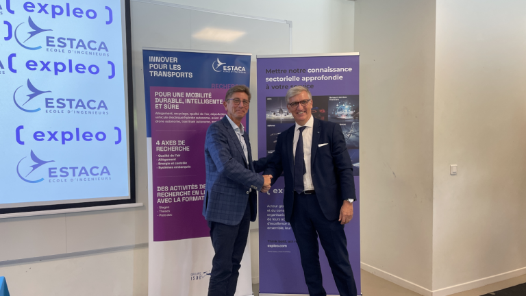 Two men in suits shake hands and smile in front of promotional banners for ESTACA and Expleo inside an office or conference room.