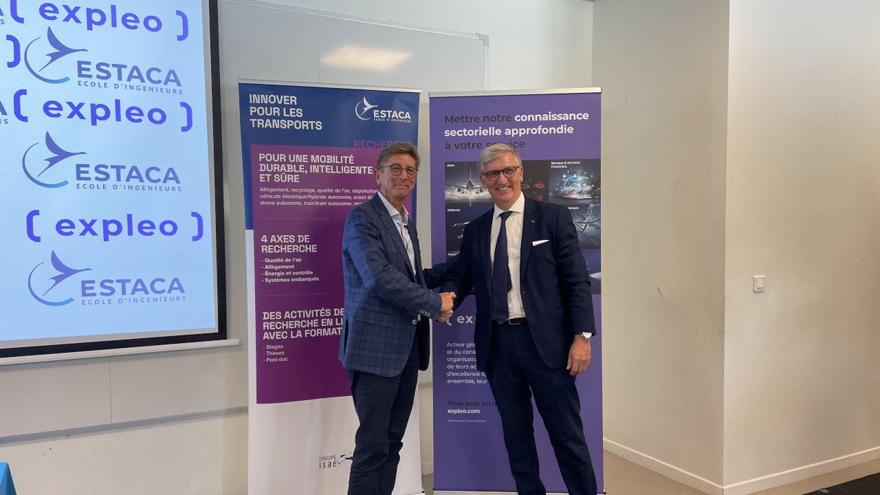 Two men in suits shake hands and smile in front of promotional banners for ESTACA and Expleo inside an office or conference room.