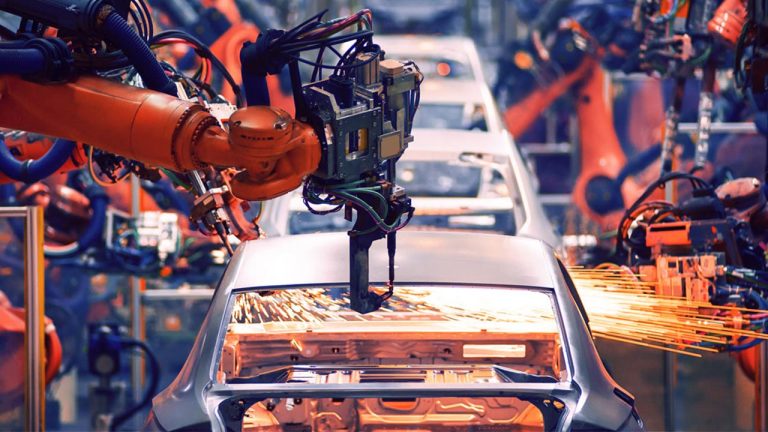 Several robotic arms work on a car body in an automobile factory, with sparks flying as one arm welds the metal. The scene is busy and industrial, showcasing automated manufacturing processes.