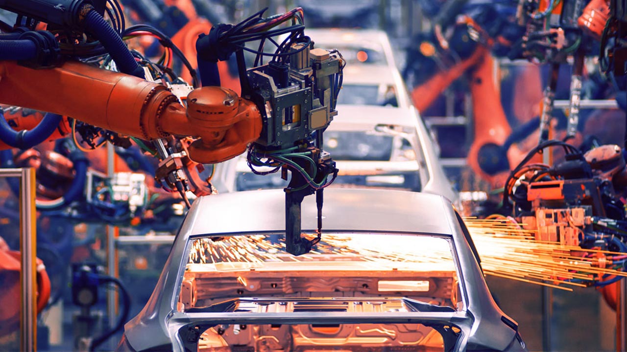 Several robotic arms work on a car body in an automobile factory, with sparks flying as one arm welds the metal. The scene is busy and industrial, showcasing automated manufacturing processes.