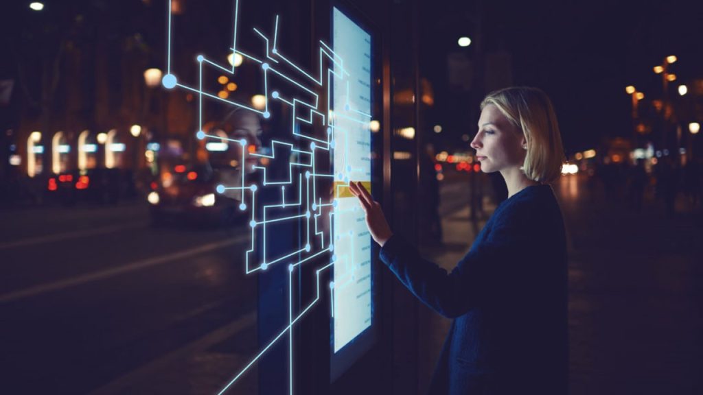A woman interacts with a digital touchscreen interface glowing with circuit-like patterns at night. The scene is set on a city street, with blurred lights and passing cars in the background.