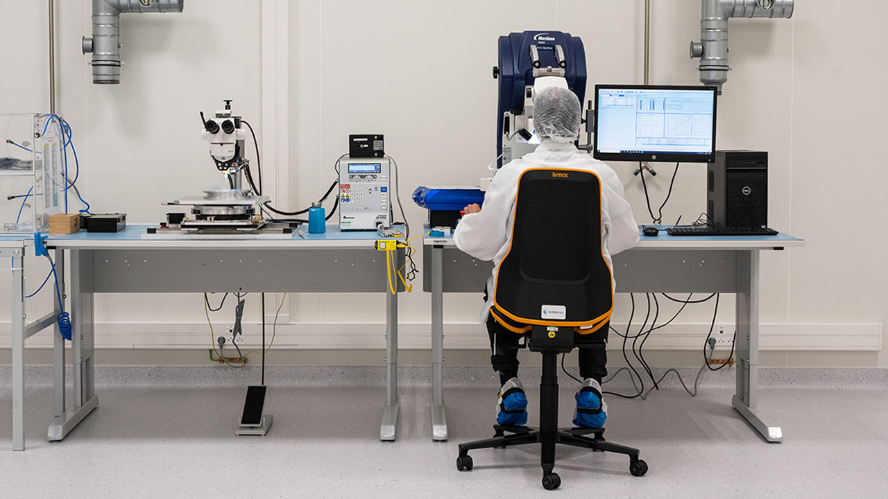 Une personne vêtue d'une combinaison de salle blanche et d'une charlotte est assise à un poste de travail équipé de matériel de laboratoire, d'ordinateurs et d'instruments scientifiques dans un environnement de laboratoire stérile.