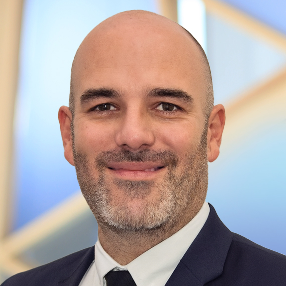 A man with a bald head and short beard, wearing a dark suit and tie, smiles at the camera. The background features a blurred, light blue and beige geometric pattern.