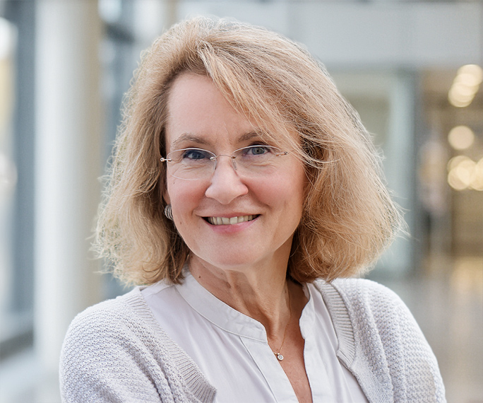 A middle-aged woman with shoulder-length blonde hair, glasses, and a light jumper smiles warmly in a bright, modern indoor setting.