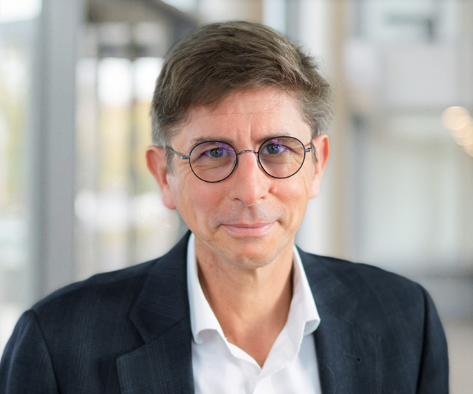 A middle-aged man with short brown hair wearing round glasses, a white shirt, and a dark blazer, standing indoors in a brightly lit, modern setting with blurred background.