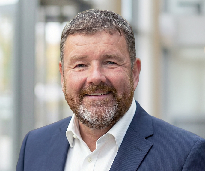 A middle-aged man with short brown hair and a beard, wearing a navy suit jacket and white collared shirt, smiling at the camera in an indoor setting with blurred background.