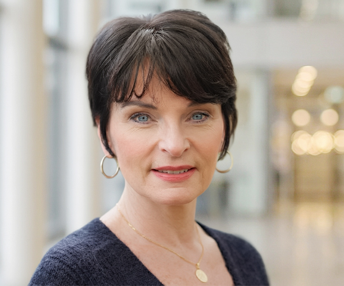 A woman with short dark hair, blue eyes, and light skin is wearing gold hoop earrings and a necklace. She is dressed in a dark jumper and standing in a brightly lit indoor setting with blurred background lights.