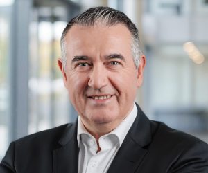A middle-aged man with short grey hair smiles confidently whilst wearing a black suit jacket and white dress shirt, standing in a modern office environment with blurred background.