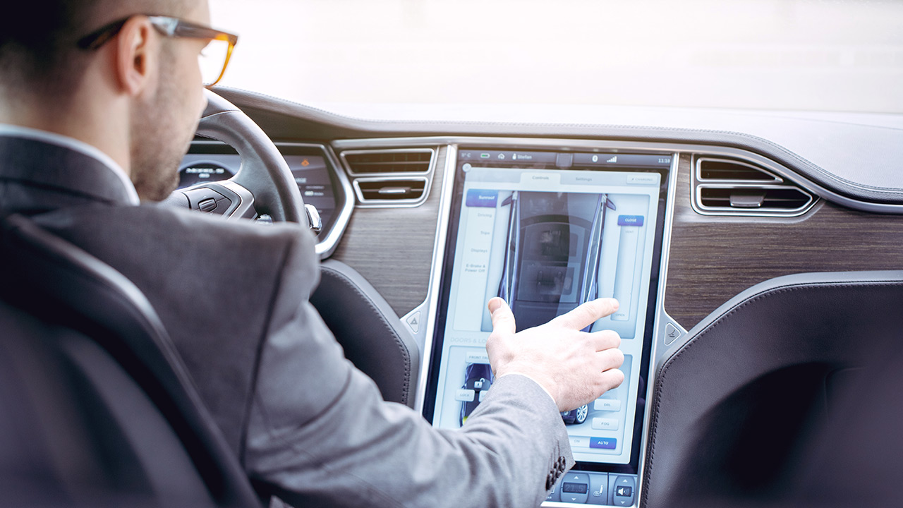 A man in a suit sits in the driver's seat of a car, using a large touchscreen display on the center console to control or view vehicle features.