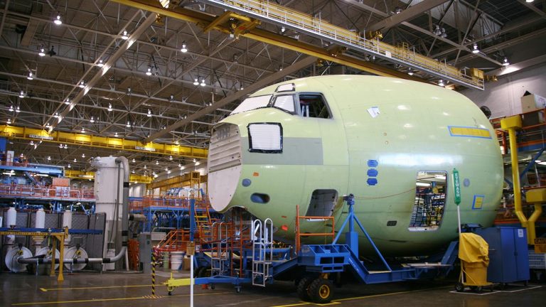 A large, unfinished airplane fuselage sits on a platform inside a spacious, well-lit aircraft manufacturing facility, surrounded by industrial equipment, scaffolding, and yellow overhead cranes.