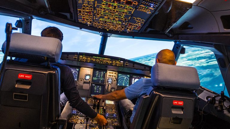 Two people sit in the cockpit of an airplane, operating the controls and instruments, with illuminated panels above and ocean scenery visible through the windshield.