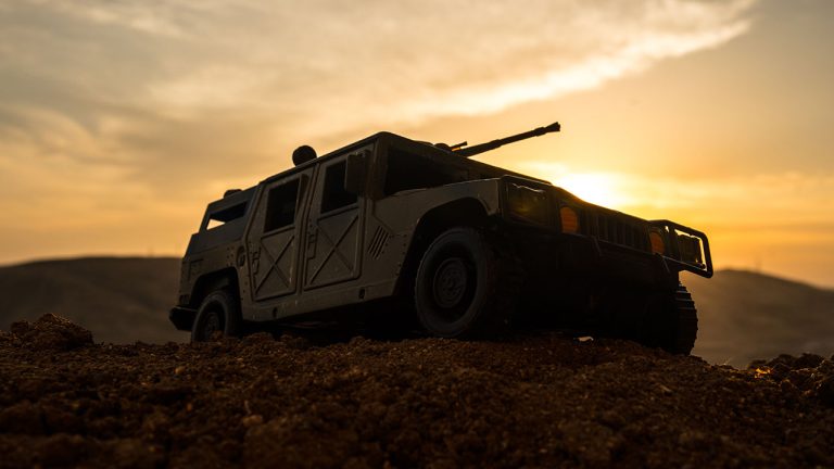 A military vehicle with a mounted gun is parked on rough terrain at sunset, silhouetted against a dramatic, cloudy sky with hills in the background.