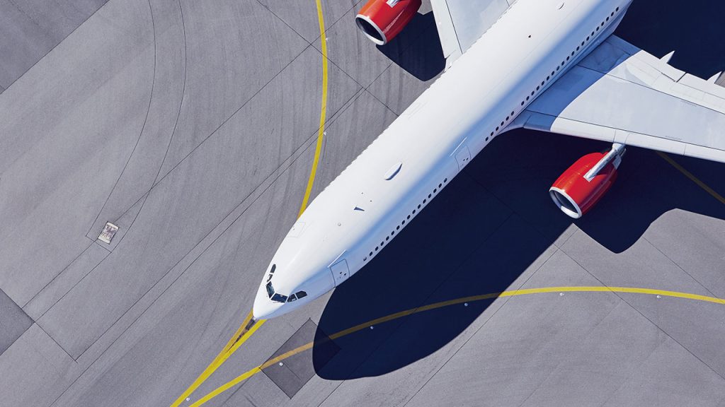Aerial view of a white airplane with red engines parked on an airport tarmac, with yellow markings visible on the gray concrete surface.