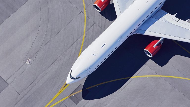 Aerial view of a white airplane with red engines parked on an airport tarmac, with yellow markings visible on the gray concrete surface.