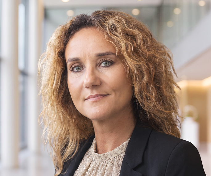 A woman with curly, light brown hair wearing a black blazer and beige top stands in a modern, bright corridor with large windows.